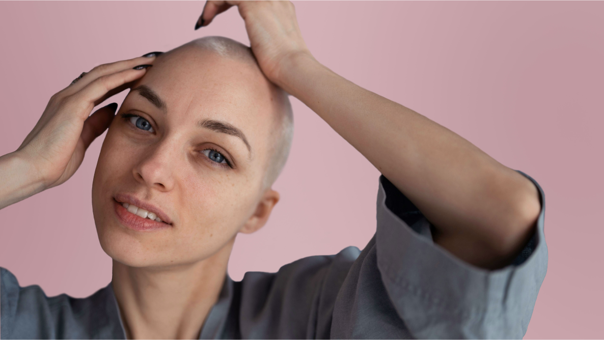 Confident woman with a shaved head touching her scalp, posing against a soft pink background.